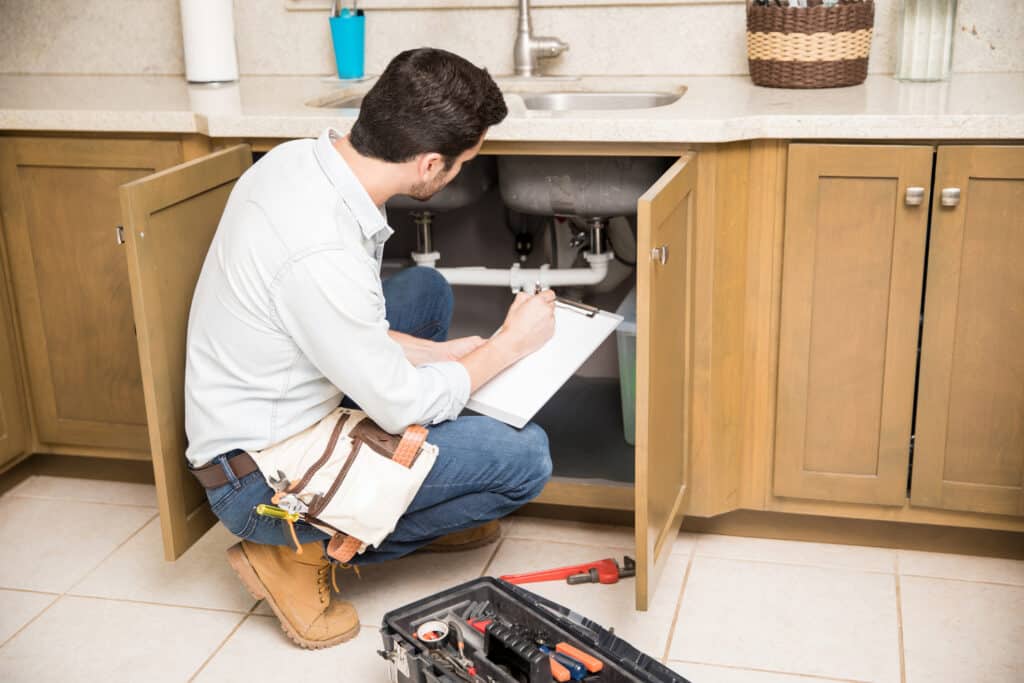 Plumber writing on a clipboard while inspecting a kitchen sink's plumbing